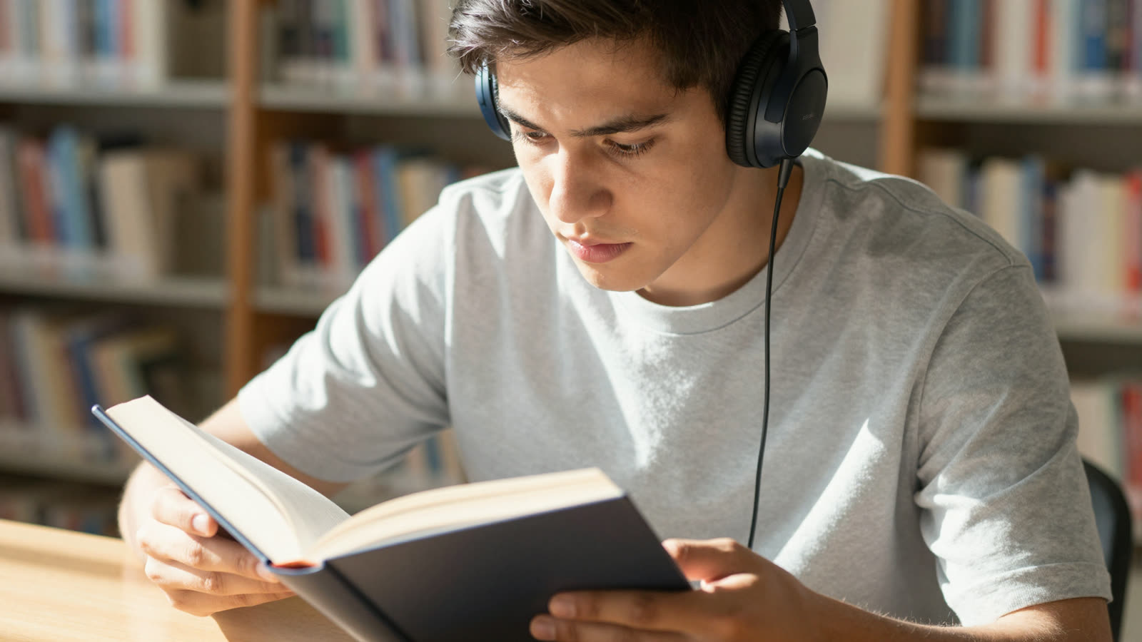 Student in a library simultaneously reading a book and listening to an audiobook.